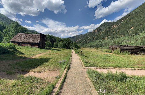 A dirt path leads through a grassy area with wooden structures and mountains under a partly cloudy sky.
