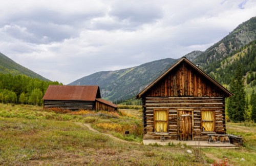Two rustic log cabins surrounded by green fields and mountains under a cloudy sky.