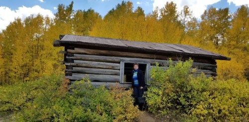A person stands by a rustic log cabin surrounded by vibrant autumn foliage and a clear blue sky.