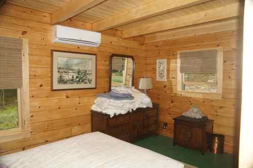 Cozy wooden bedroom with a bed, dresser, mirror, lamp, and air conditioning unit. Natural light from windows.