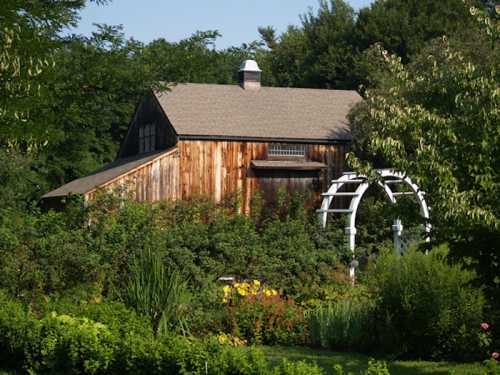 A rustic wooden barn surrounded by lush greenery and colorful flowers under a clear blue sky.