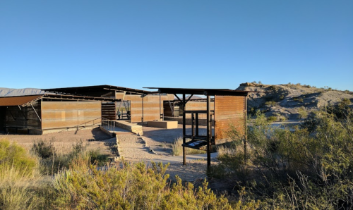 Modern building in a desert landscape, featuring metal and wood materials, surrounded by sparse vegetation and hills.