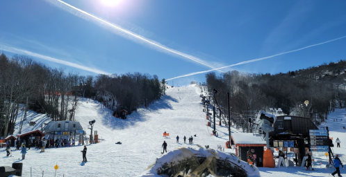 A sunny ski slope with people skiing, snow-covered trees, and a clear blue sky with contrails above.
