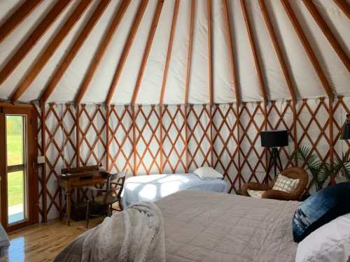 Interior of a yurt featuring wooden beams, a bed, a chair, a desk, and large windows with natural light.