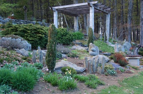A serene garden with stone sculptures, lush greenery, and a wooden pergola surrounded by trees.