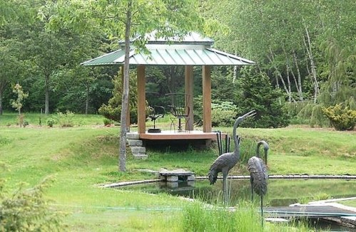 A serene garden scene featuring a gazebo, pond, and decorative cranes amidst lush greenery.