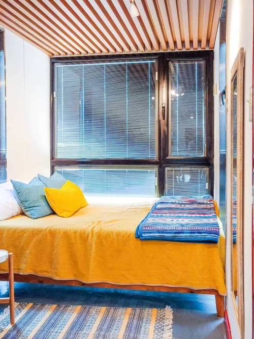 Cozy bedroom with a yellow bedspread, colorful pillows, and large windows letting in natural light.