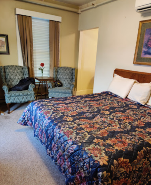 Cozy bedroom with a floral quilt, two armchairs, a small table, and a window with curtains.