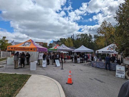 A lively outdoor market scene with colorful tents, people browsing, and a clear blue sky with clouds.