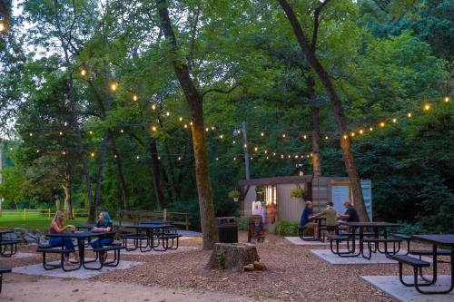 A cozy outdoor gathering space with picnic tables, string lights, and people socializing among trees.