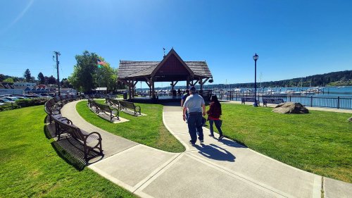 A couple walks along a path in a park by the water, with a gazebo and boats in the background under a clear blue sky.