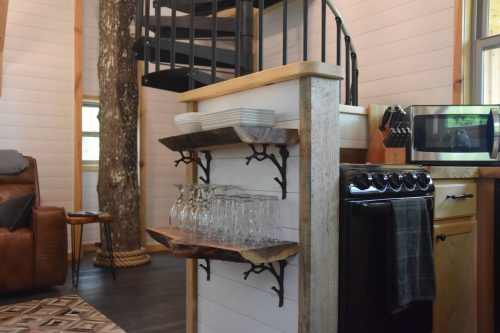 Cozy interior of a cabin featuring a spiral staircase, shelves with glasses, and a kitchen area with a stove and microwave.