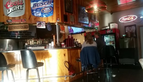 A cozy bar interior with wooden walls, two patrons sitting at the counter, and various beer signs on display.