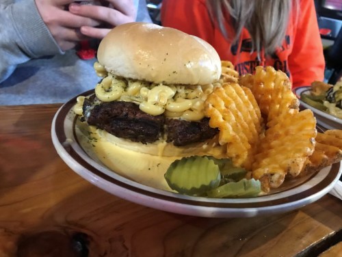 A close-up of a burger with macaroni and cheese on a bun, served with waffle fries and pickles on a plate.