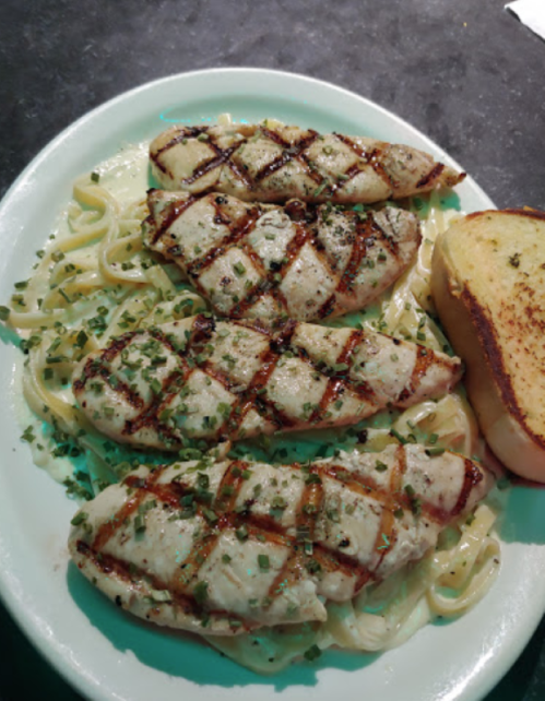 Grilled chicken breasts on a bed of fettuccine, garnished with herbs, served with a slice of toasted bread.