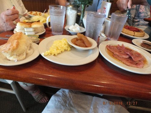 A table set with breakfast plates: biscuits, scrambled eggs, ham, apple slices, and a waffle with syrup.
