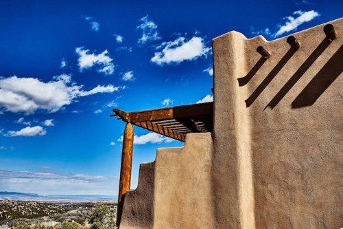 A southwestern-style adobe structure with a wooden pergola against a bright blue sky and distant landscape.