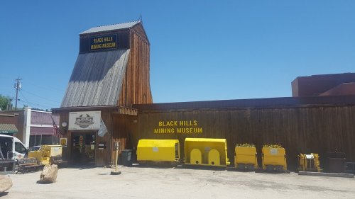 Exterior of the Black Hills Mining Museum, featuring a wooden building and yellow mining equipment in front.