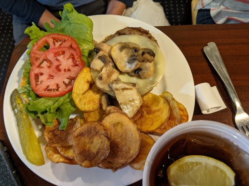A plate with a burger topped with mushrooms and onions, lettuce, tomato, potato chips, a pickle, and a drink with lemon.