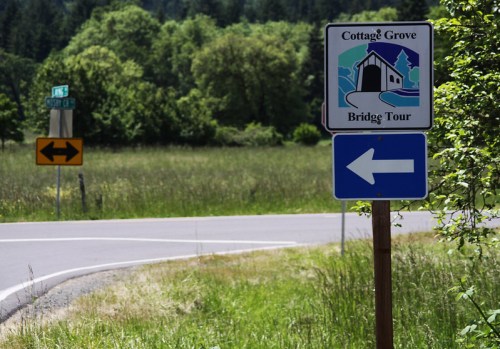 Sign for Cottage Grove Bridge Tour with directional arrows, set against a green landscape and road.
