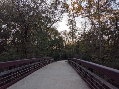 A wooden bridge leads into a wooded area, with trees and a soft sunset glow in the background.