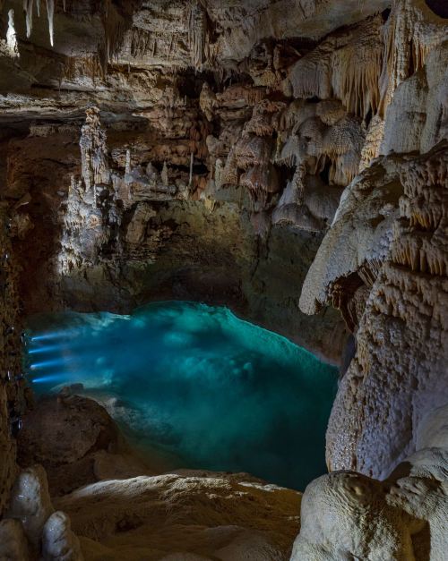 A stunning underground cave with stalactites and a glowing turquoise pool of water illuminated by soft light.