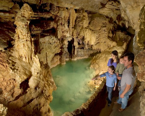 A family explores a cave with stalactites and a turquoise pool, marveling at the natural formations around them.