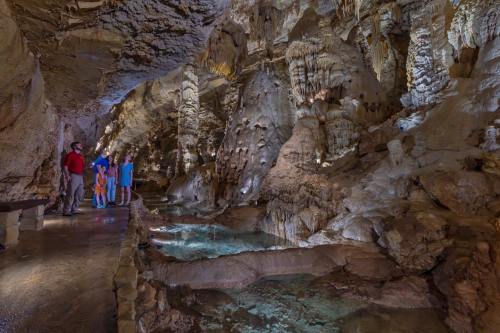 A group of visitors explores a cavern with stunning rock formations and water features, illuminated by soft lighting.