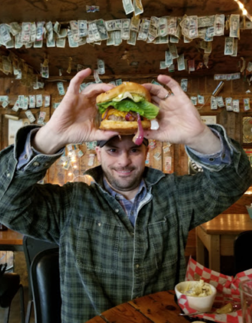 A person holds up a large burger in a rustic restaurant decorated with dollar bills.