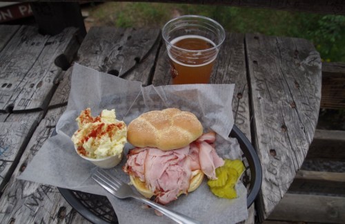 A plate with a sandwich filled with ham, a side of potato salad, a pickle, and a cup of beer on a wooden table.