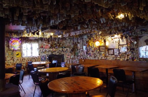 A rustic bar interior with wooden tables, black chairs, and walls and ceiling covered in dollar bills and memorabilia.
