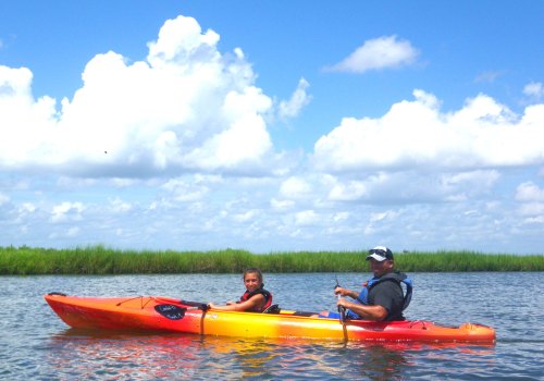 A man and a child paddle a kayak on a calm waterway under a blue sky with fluffy clouds and green grass in the background.