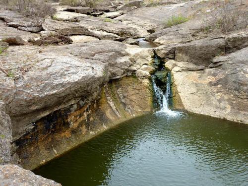A small waterfall cascades into a tranquil pool surrounded by rocky terrain and sparse vegetation.