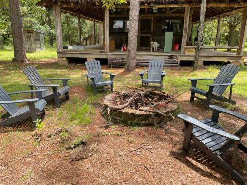 A circular fire pit surrounded by black Adirondack chairs, with a rustic cabin in the background and trees around.