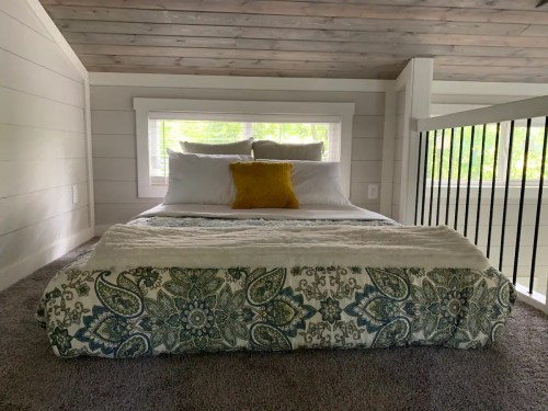 Cozy loft bedroom with a bed adorned in white linens and a patterned blanket, featuring a yellow pillow and natural light.