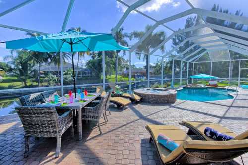 A sunny poolside area with lounge chairs, a table, and umbrellas, surrounded by greenery and a sparkling pool.