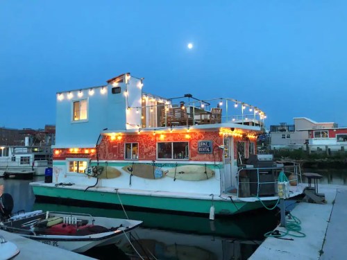 A colorful houseboat illuminated with string lights, docked at a marina under a moonlit sky.