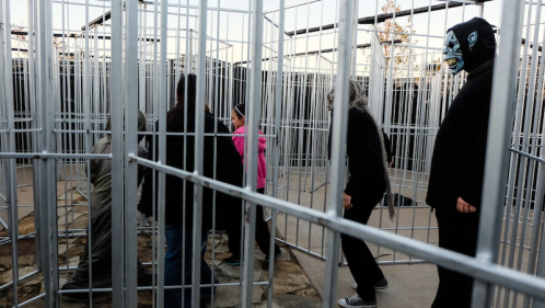 A group of people navigate a maze-like structure made of metal bars, with one person wearing a mask in the background.