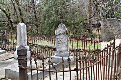 A historic cemetery with old gravestones surrounded by a wrought iron fence and overgrown trees in the background.