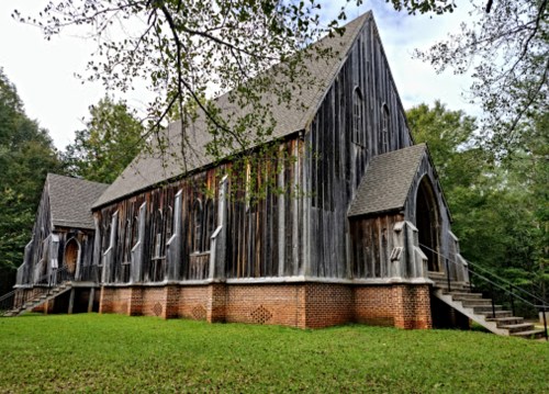 A rustic wooden church with a steep roof, surrounded by trees and a grassy area, featuring brick accents and steps.