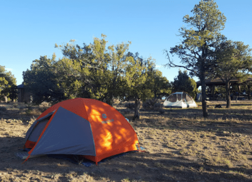 A bright orange tent set up in a grassy campsite surrounded by trees and another tent in the background.