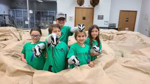 A group of five children in green shirts hold toy animals, standing on a sandy surface with a natural setting backdrop.