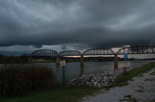 A dark, cloudy sky looms over a river with a metal bridge in the background and rocky shoreline in the foreground.