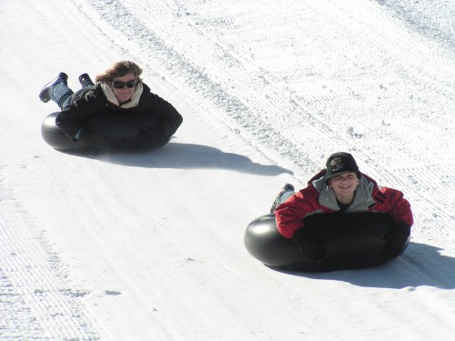 Two people sledding down a snowy hill on black inner tubes, smiling and enjoying the winter fun.