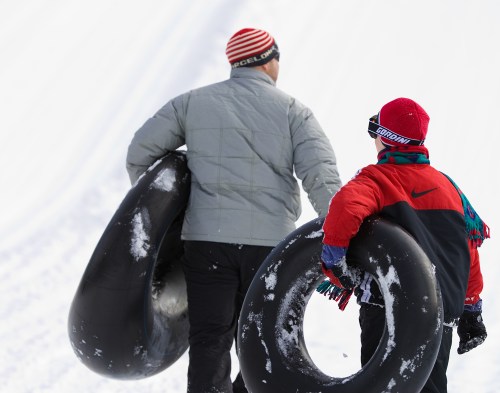 Two people walk up a snowy hill, each carrying a black snow tube, dressed in winter jackets and hats.
