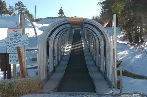 A snow-covered sledding ramp with a covered conveyor belt, surrounded by trees and a warning sign.