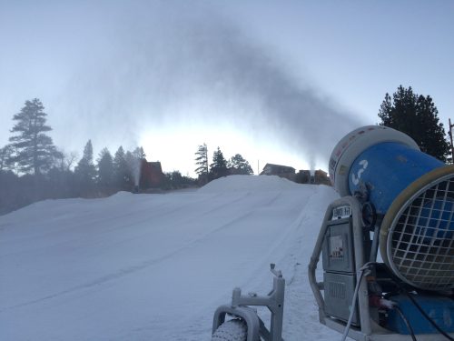 A snowmaking machine sprays artificial snow on a ski slope, with trees and buildings in the background.