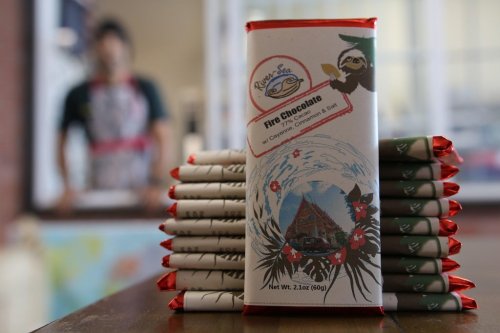 A stack of chocolate bars with tropical designs, featuring a label that reads "Fire Chocolate." A person is blurred in the background.