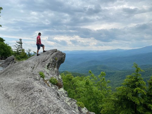 A person stands on a rocky outcrop, overlooking a lush green valley and distant mountains under a cloudy sky.