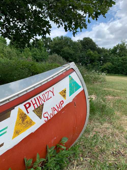 A colorful canoe with "Phinizy Swamp" painted on it, set against a backdrop of green trees and a cloudy sky.